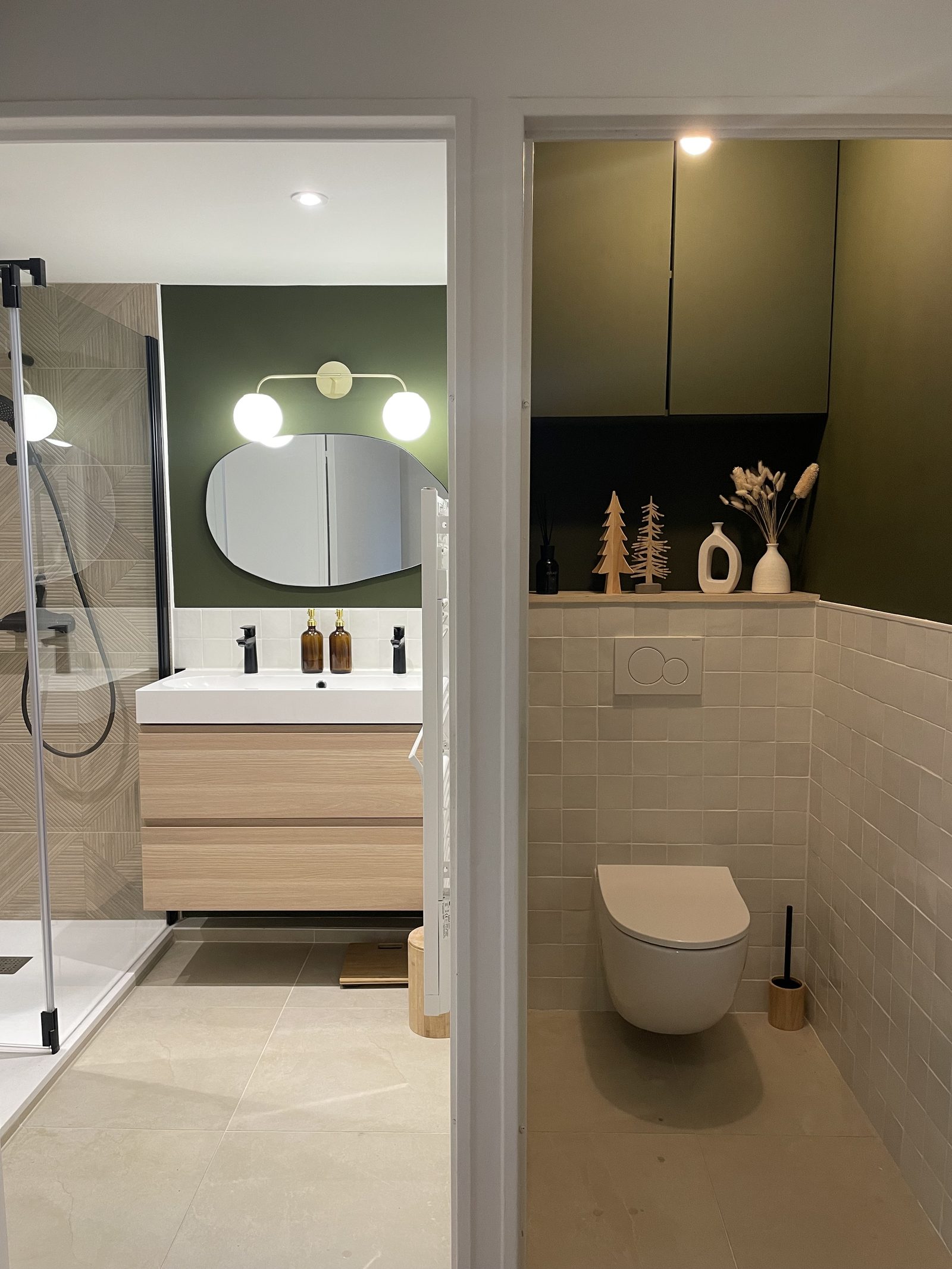 Vue d'ensemble de la salle de bains rénovée : meuble vasque bois, mur vert olive, miroir ovale et coin WC à Lyon